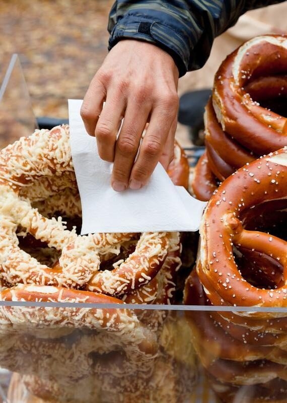 A hand holding a white napkin reaches for a cheese-topped pretzel next to stacks of salted pretzels behind glass.