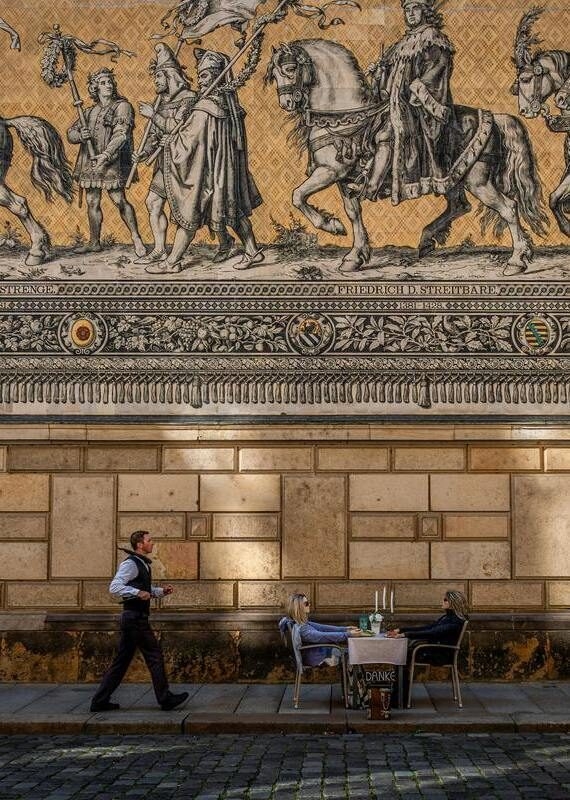 People dine at a sidewalk table beneath the massive Procession of Princes mural in Dresden.