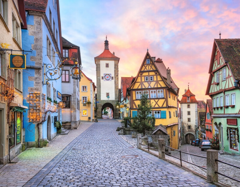 A cobblestone street splits between colorful half-timbered buildings and stone towers under a sunset sky.