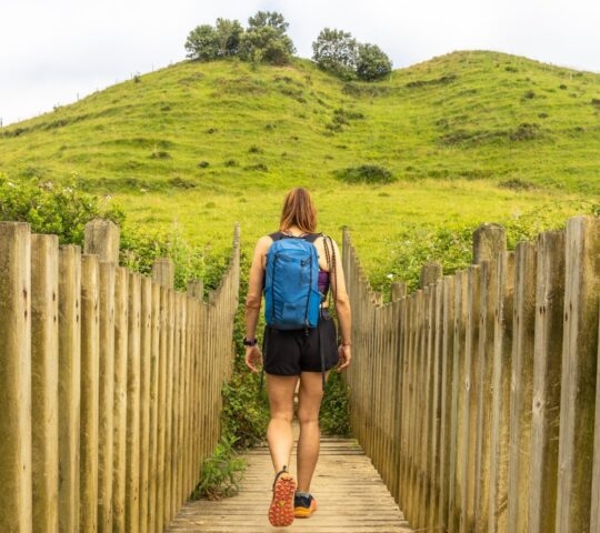 A hiker with a blue backpack walking on a wooden path lined with posts towards a green hill on the Camino de Santiago.