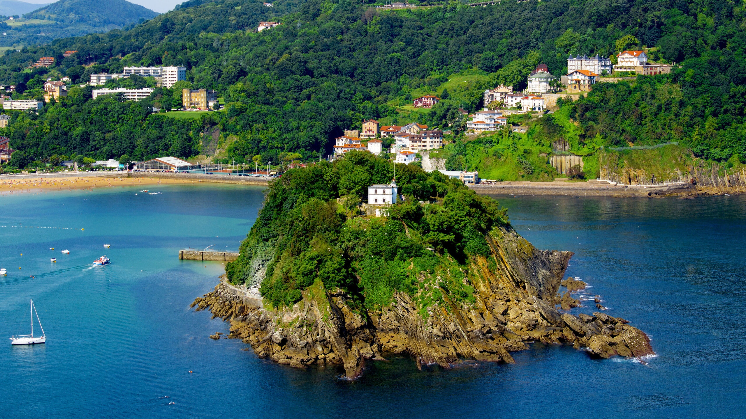 A rocky, tree-covered island in a blue bay, surrounded by boats and a hilly, populated coastline. Keyword: kayaking.