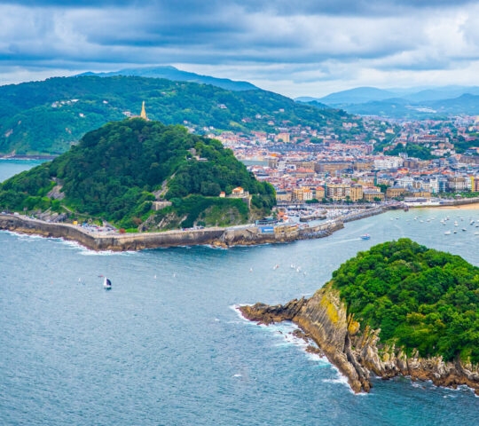 A rocky, tree-covered island in a blue bay, surrounded by boats and a hilly, populated coastline. Keyword: kayaking.