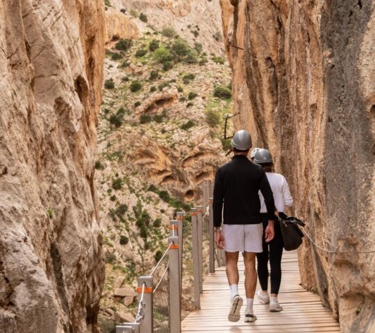 Two people in helmets on a private hike to el caminito del rey malaga, walking on a wooden cliffside boardwalk.