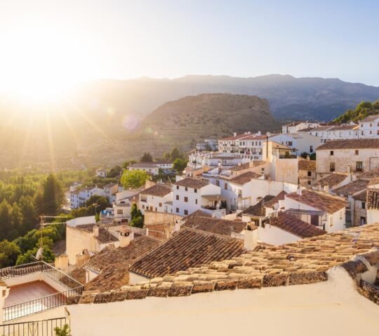 Sun setting over a pueblo blanco andaluz, showing white houses with terracotta roofs nestled in a green mountain valley.