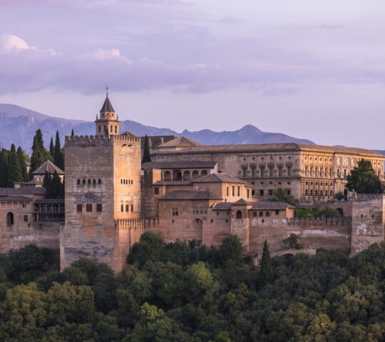 A large stone fortress and palace complex on a wooded hill with mountains in the background under a purple evening sky.