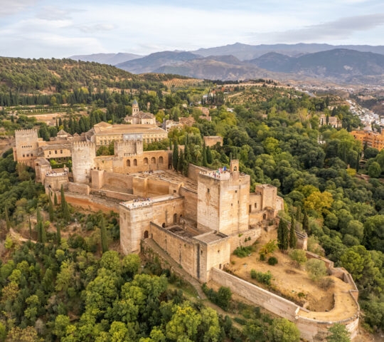 Aerial view of the Alhambra palace and fortress complex in Granada, Spain, set on a forested hill near mountains.