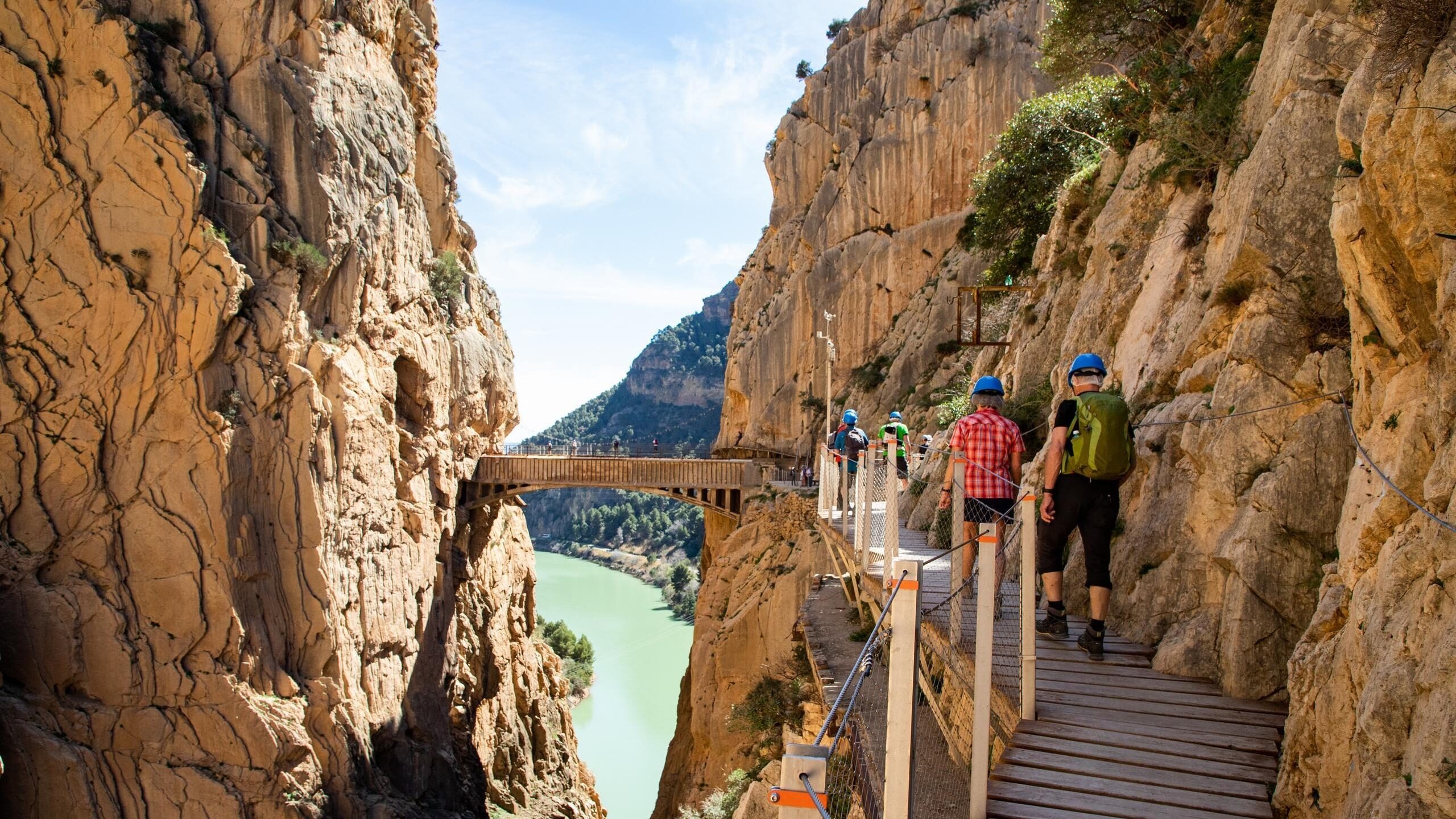 Hikers in helmets walk along a narrow wooden boardwalk attached to a steep rocky cliff over a river at Caminito del Rey.