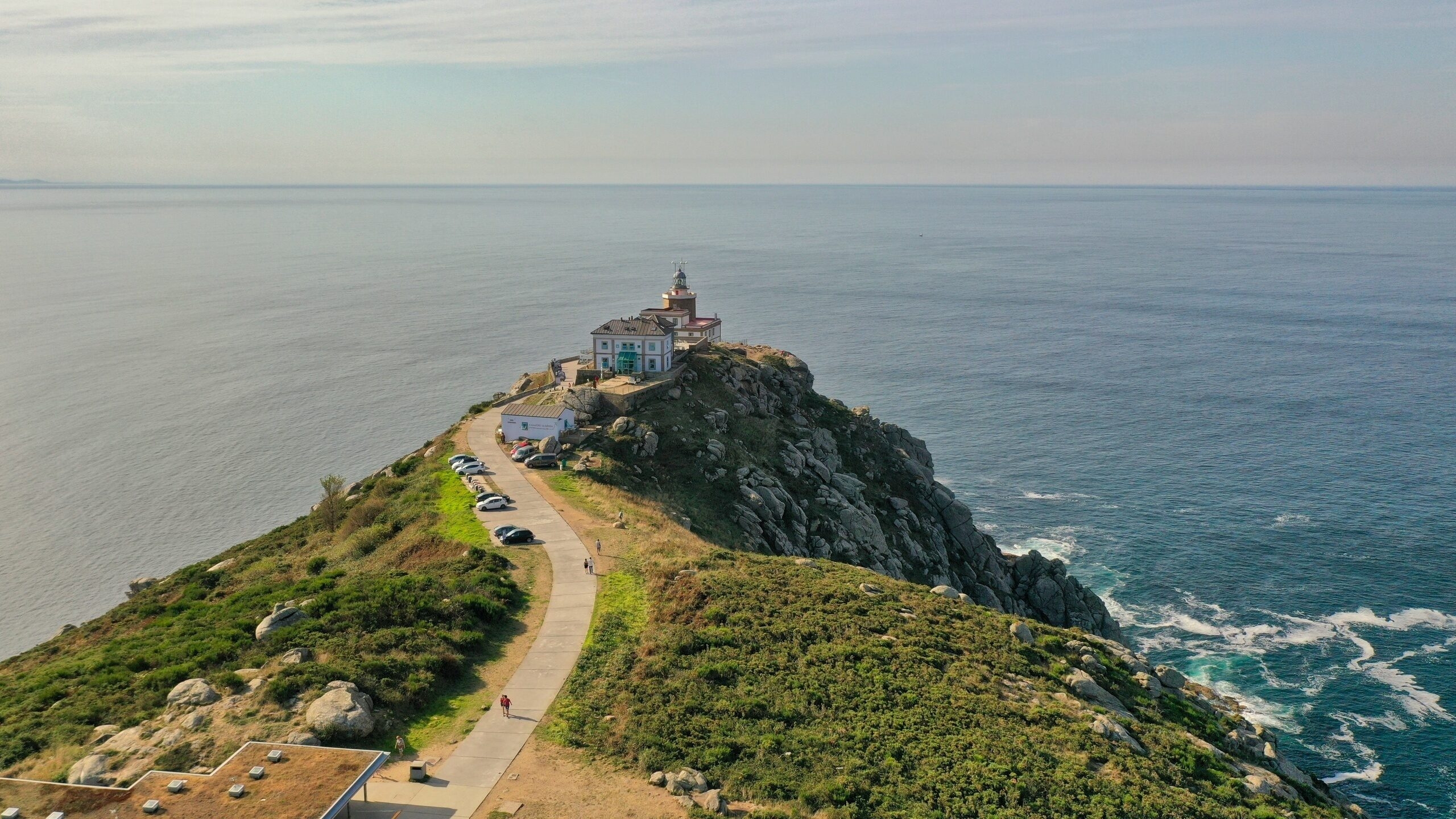 Aerial view of a lighthouse at the end of a winding paved road on a steep, rocky green cape extending into the ocean.