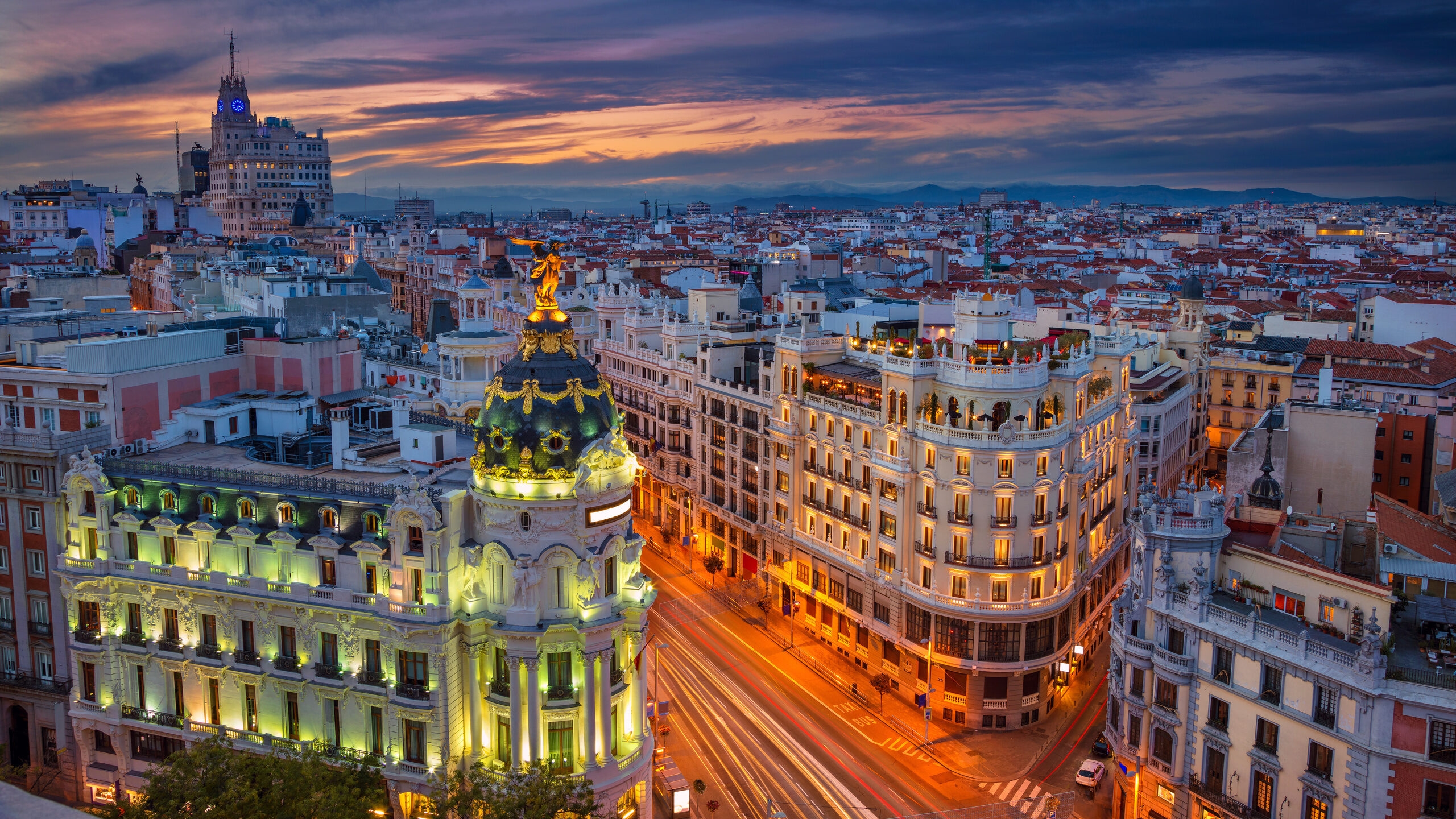 Elevated view of the illuminated Metropolis building and busy city streets in Madrid at dusk under a colorful sky.