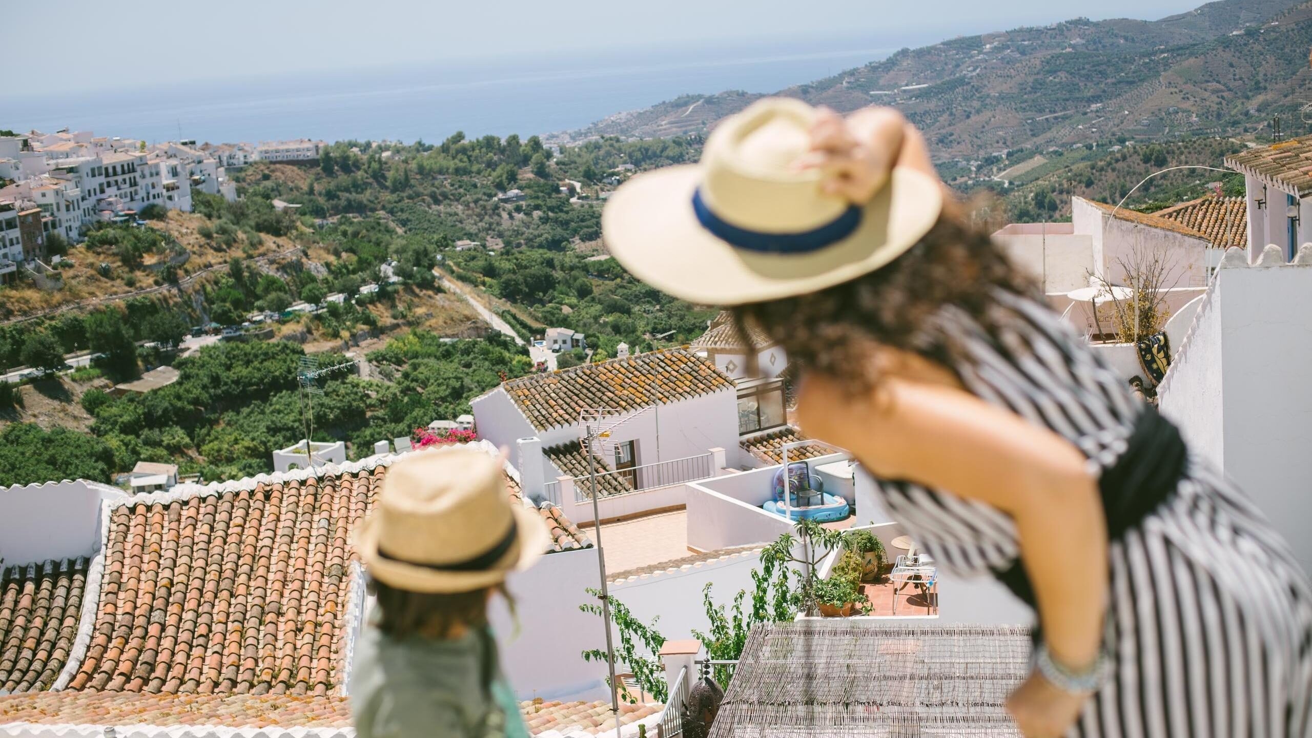 Two people wearing straw hats look out over a hillside traditional white village and mountainous landscape in Malaga.