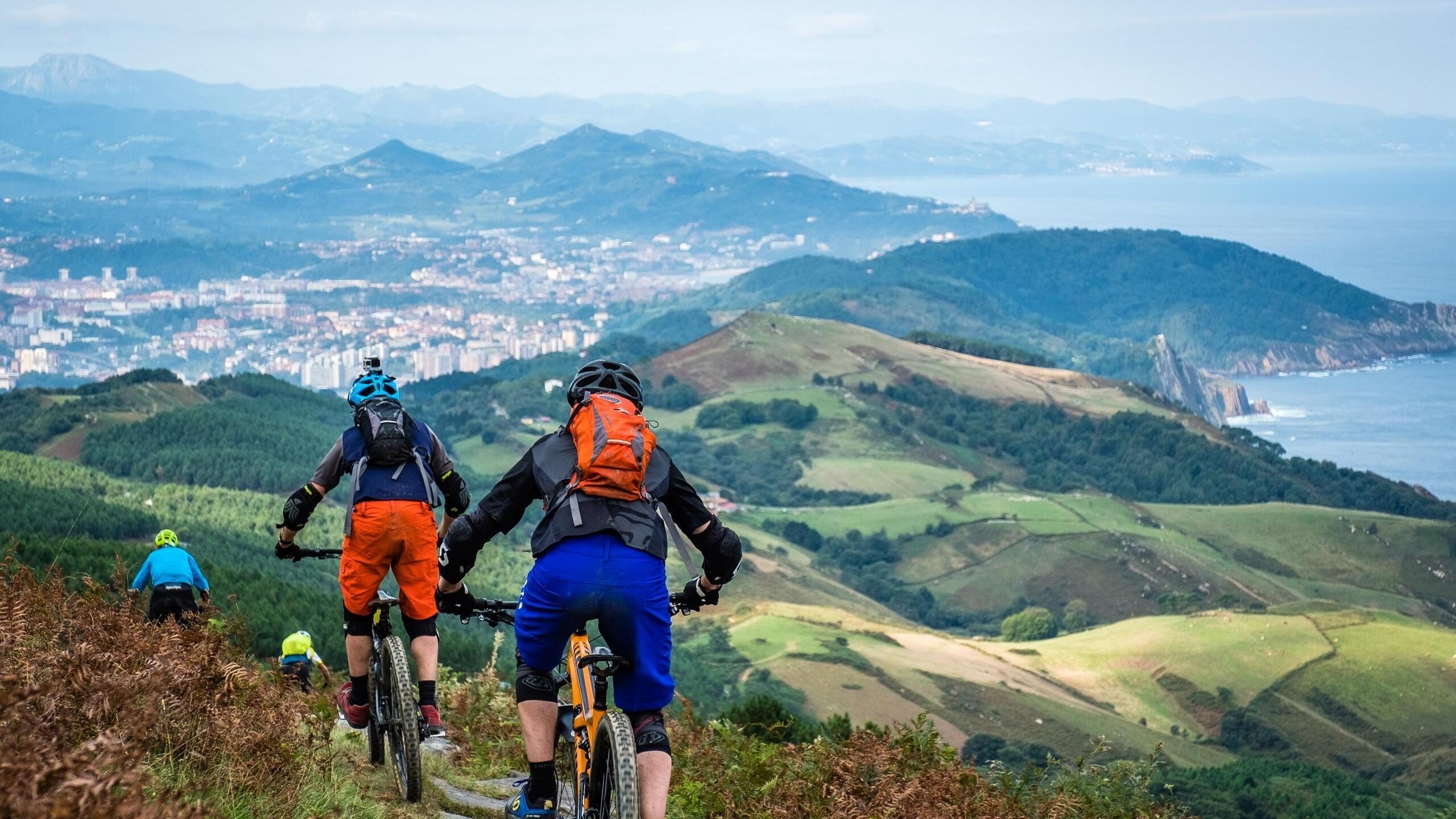 Mountain bikers with backpacks riding a hillside trail overlooking the coastal city of San Sebastian and the ocean.