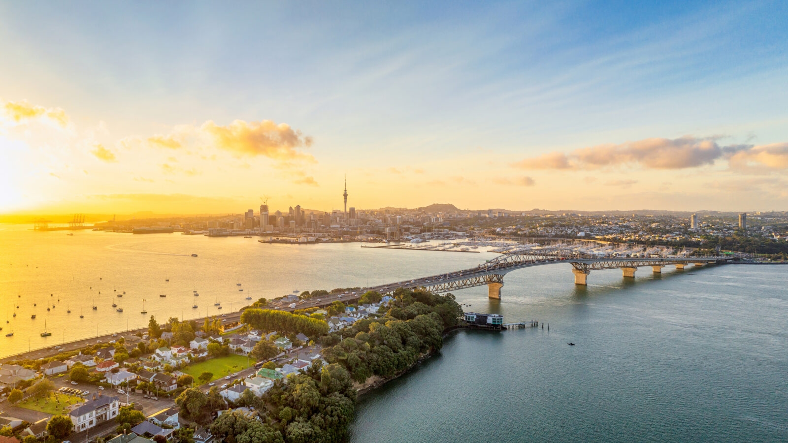 Aerial view of the Auckland skyline and Harbour Bridge at sunset during a family-trip, with boats on the golden water.