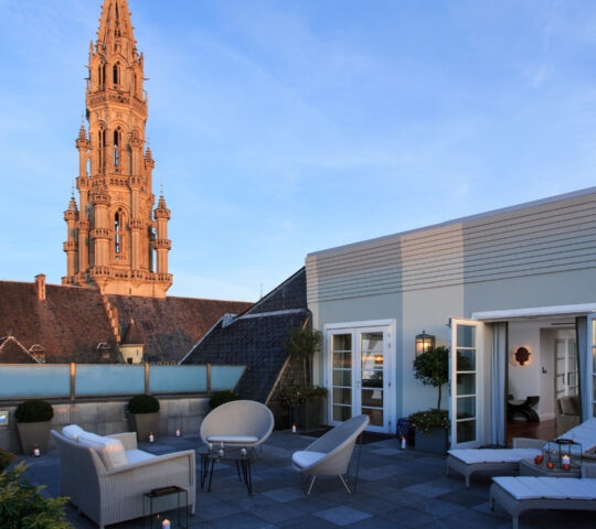 Rooftop terrace at Hotel Amigo Belgium with lounge seating, candles, and views of a tall gothic spire at dusk.