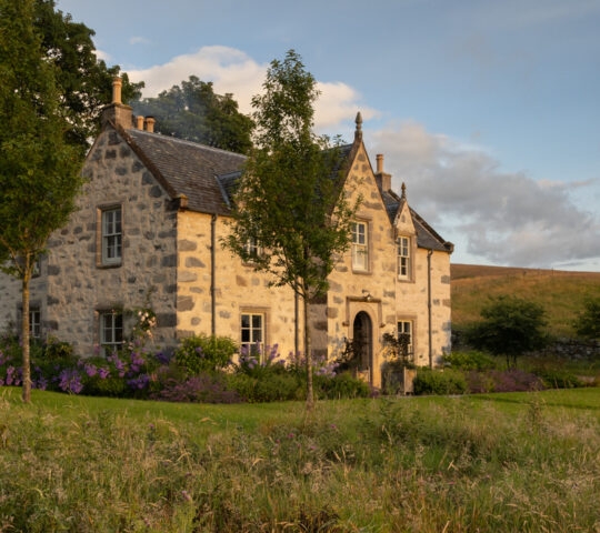 Stone farmhouse in mid distance with green meadow in the foreground and Scottish fells and blue sky beyond.