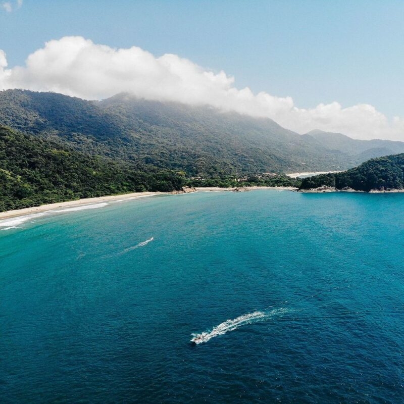 Aerial view of a boat in blue water near a sandy beach and green forested mountains with low clouds in Costa Verde, Brazil.