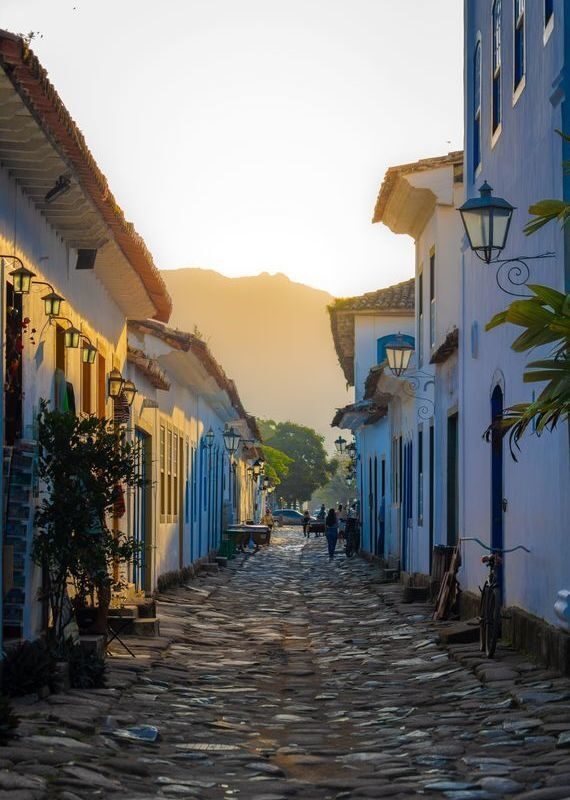 A cobblestone street lined with white buildings featuring blue trim, lit by warm sunlight in Paraty, Brazil.