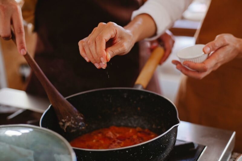 Two people in aprons cooking in Brazil; one stirs a red sauce in a pot while the other sprinkles salt from a small bowl.
