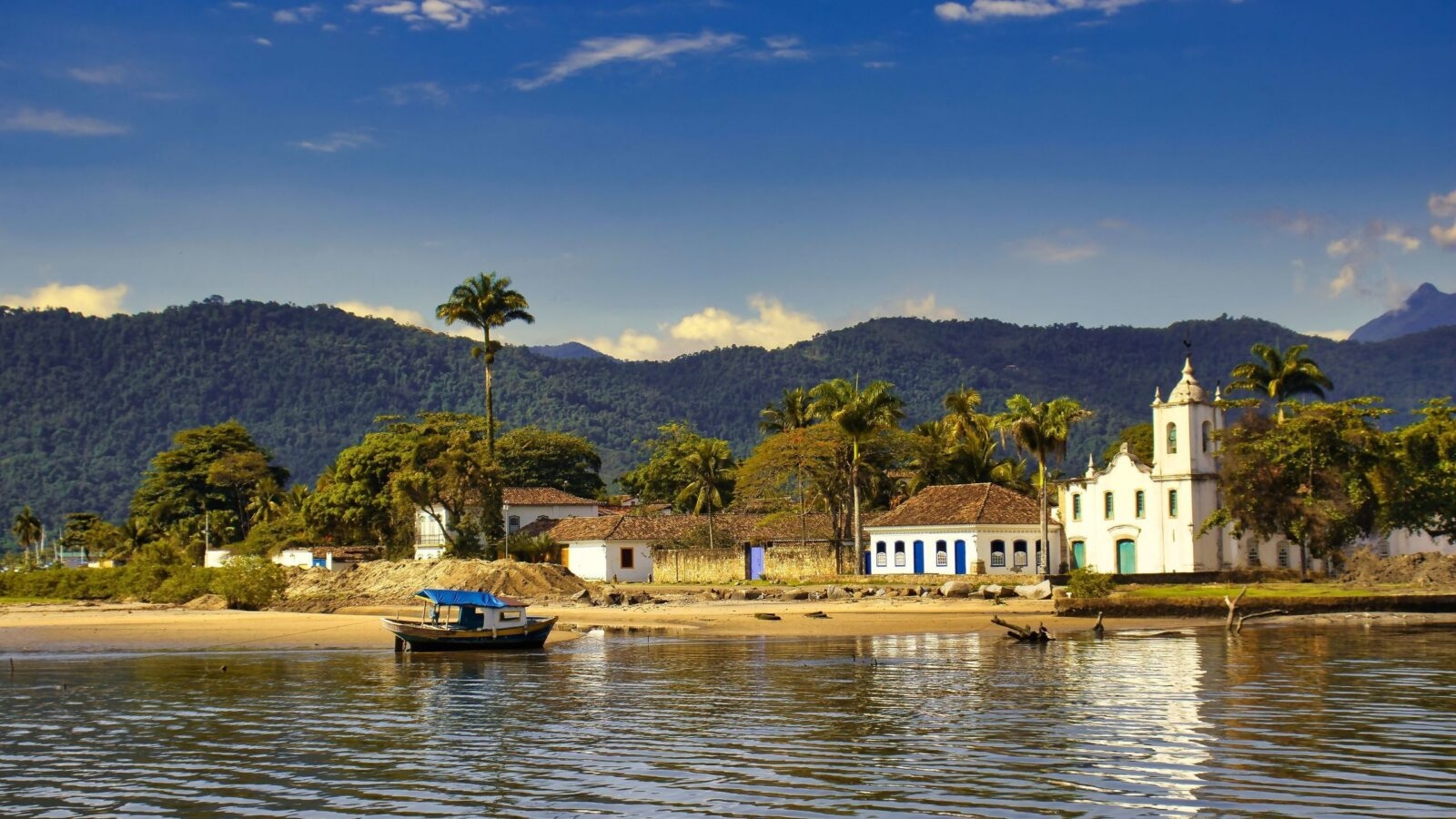 View from the water of white colonial buildings, a church, and a small boat on the shore in Paraty, Brazil.