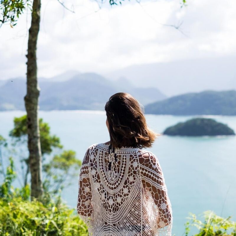 A woman seen from behind wearing a white crocheted top, looking at a bay with islands and mountains in Picinguaba, Brazil.