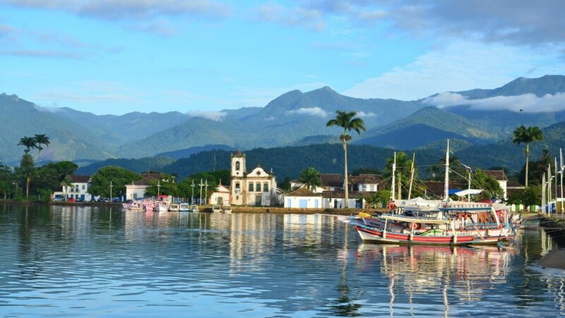 Boats moored in the water before the historic white buildings and green mountains of Paraty, Brazil.