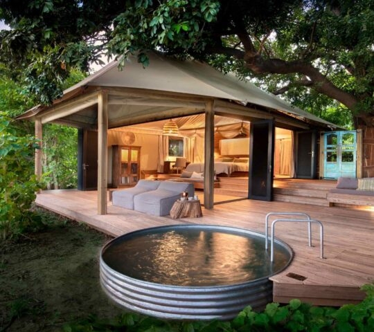 A view of a wooden deck with a plunge pool and a luxury suite behind. The sides of the tented room are open to the outside.