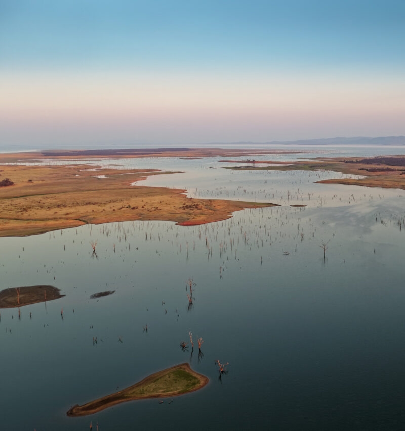Aerial view of Lake Kariba at sunset, showing a peninsula with dry vegetation and submerged tree trunks in calm water.