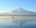 A small boat on a misty lake reflecting a large snow-capped mountain under a clear blue sky.