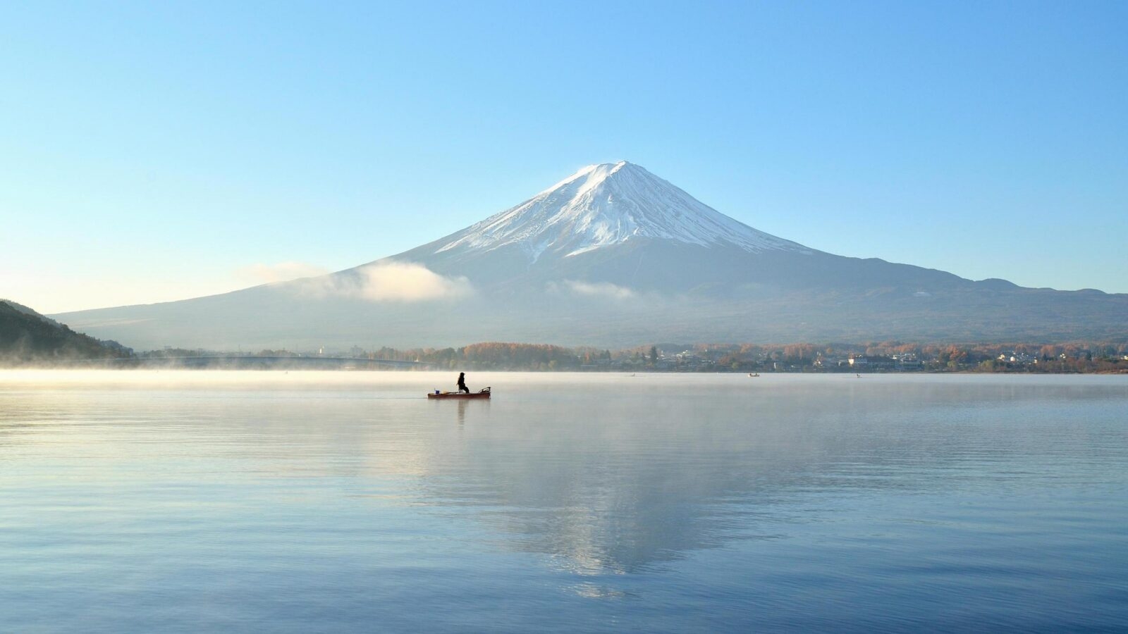 A small boat on a misty lake reflecting a large snow-capped mountain under a clear blue sky.