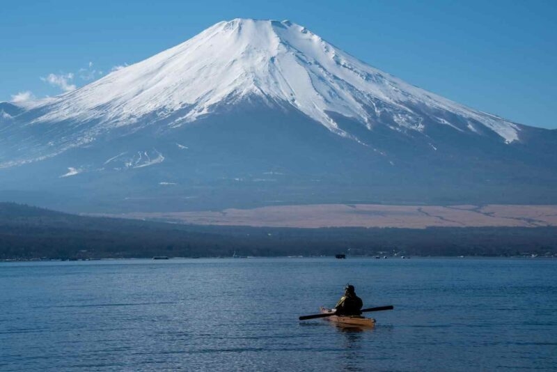 Kayaker on a calm lake in front of a massive snow-capped mountain under a clear blue sky.