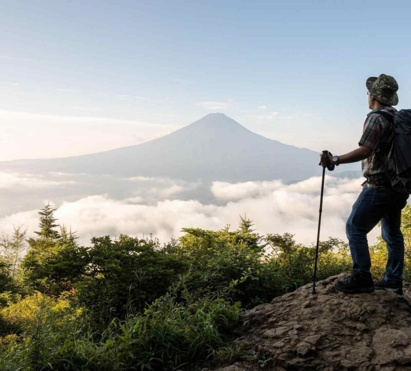 Hiker with a backpack and trekking pole standing on a rock looking at a distant mountain peak above clouds.