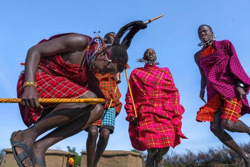 Maasai men wearing red plaid garments performing a jumping dance outdoors under a bright blue sky.