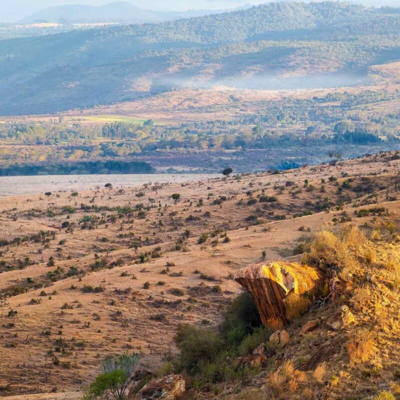 Sunlit rock formation overlooking a wide dry valley with rolling hills in the background under soft daylight.
