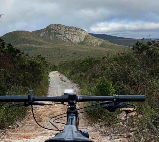 First-person view over mountain bike handlebars on a dirt trail facing a mountain.